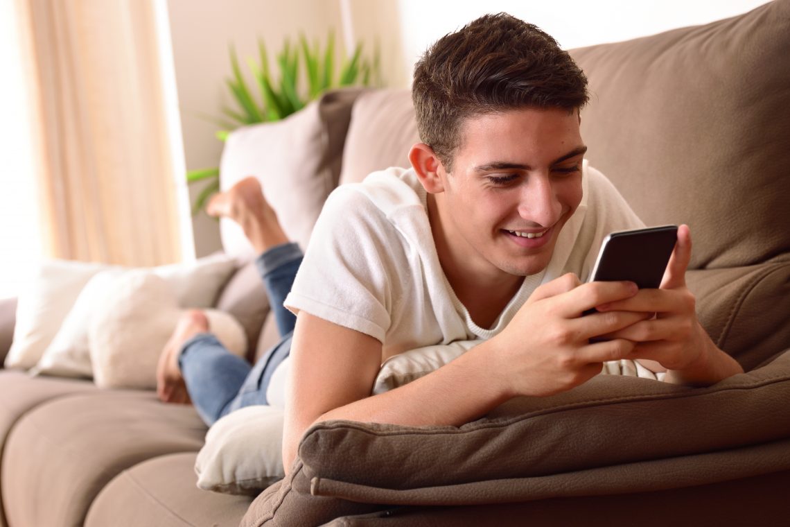 Ein junger Mann liegt auf einem Sofa und schaut auf sein Telefon.